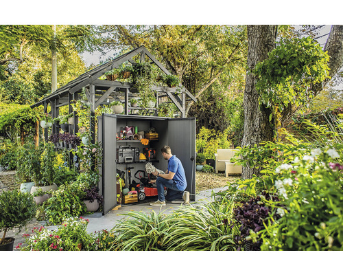 Scéna so záhradnou kôlňou plnou záhradného náradia a vybavenia, obklopenou bujnou zeleňou