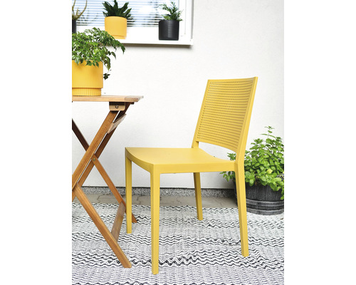 Žltá plastová záhradná stolička s perforovaným operadlom vedľa dreveného stola na vzorovanom koberci