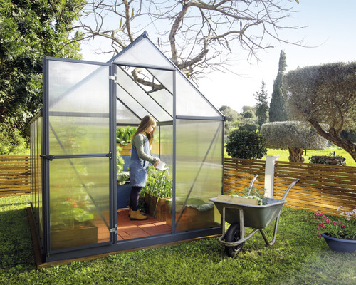 Skleník s otvorenými dverami v záhrade, žena zalieva rastliny.