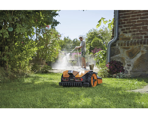 Robotická kosačka na trávniku, v pozadí osoba čistí terasu vysokotlakovým čističom