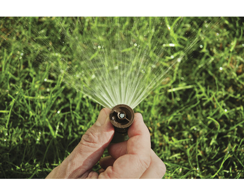 Ruka drží zavlažovač trávnika, ktorý strieka vodu.