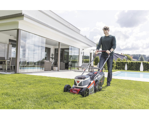 Muž kosí trávnik akumulátorovou kosačkou v záhrade s bazénom.