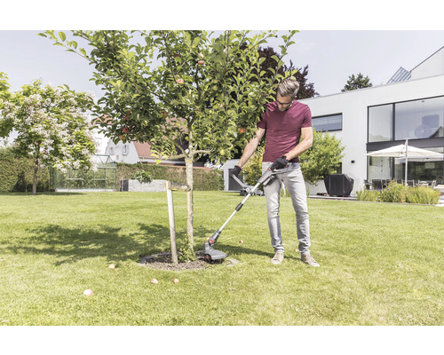 Muž zastrihuje trávnik strunovou kosačkou okolo stromu v záhrade.