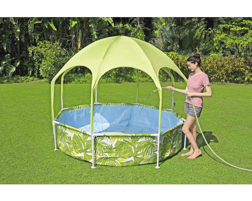 Záhradná scéna s bazénom s rámovou konštrukciou a vzorom listov a slnečnou strieškou, zatiaľ čo žena napúšťa bazén záhradnou hadicou.