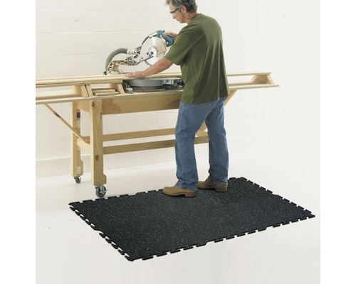 Osoba obsluhuje pokosovú pílu na pracovnom stole a stojí na čiernej dielňovej podložke.