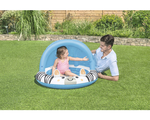 Dieťa v bazéniku so zebrovým vzorom a slnečnou ochranou, dospelý leží vedľa na trávniku