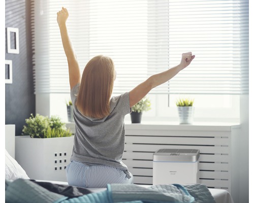 Žena sedí na posteli, naťahuje sa a pozerá sa na čističku vzduchu v spálni.