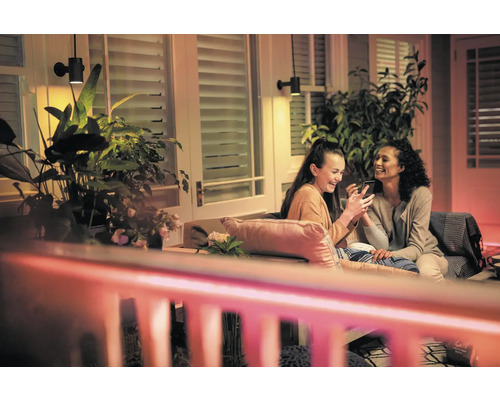 Scéna s dvoma ženami sediacimi na terase pod vonkajšími nástennými svietidlami, ktoré sa pozerajú na smartfón, obklopené rastlinami.