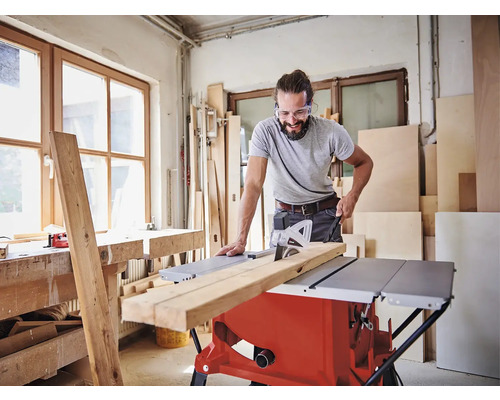 Muž s ochrannými okuliarmi reže drevenú dosku stolovou pílou v dielni.