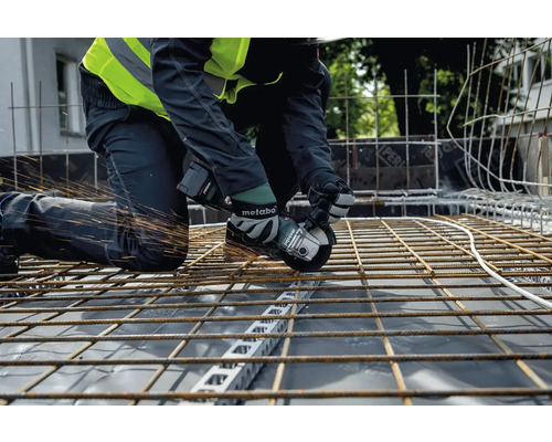 Remeselník kľačí na stavenisku a reže oceľové nosníky uhlovou brúskou Metabo, pričom lietajú iskry.