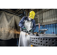 Pracovník s ochrannou prilbou, chráničmi sluchu, respirátorom a zásterou brúsi kov uhlovou brúskou, pričom lietajú iskry.