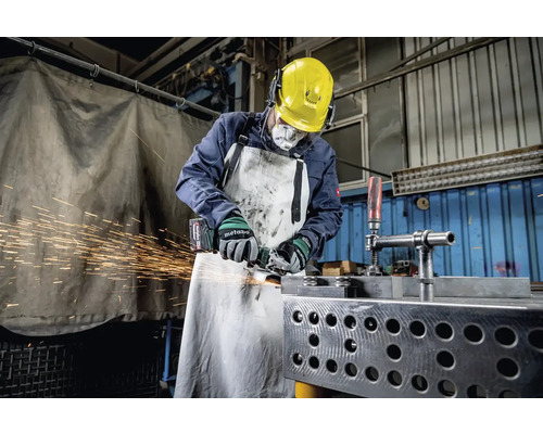 Pracovník s ochrannou prilbou, chráničmi sluchu, respirátorom a zásterou brúsi kov uhlovou brúskou, pričom lietajú iskry.