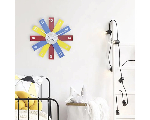 Scéna s nástennými hodinami s farebnými ciferníkmi, rebríkom so žiarovkami a posteľou.