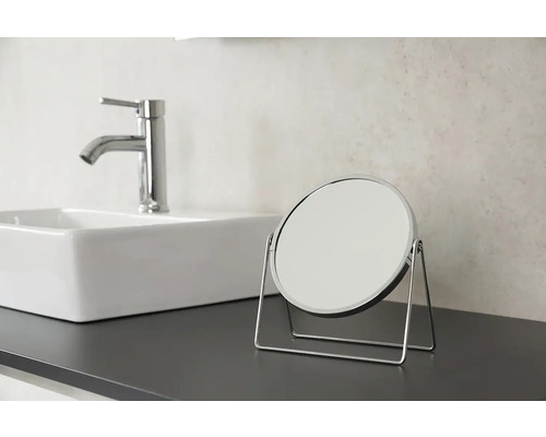 Okrúhle kozmetické zrkadlo s chrómovaným rámom na doske umývadla vedľa umývadla a batérie.