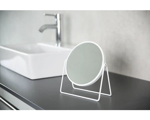 Okrúhle kozmetické zrkadlo s kovovým rámom na kúpeľňovej poličke s umývadlom a vodovodnou batériou v pozadí.