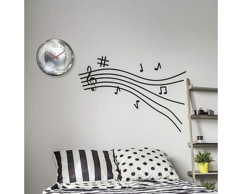 Nástenná dekorácia so zrkadlovými hodinami a nálepkou na stenu s notovými linkami nad posteľou.