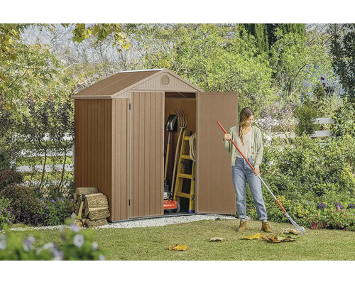 Záhradná scéna s kôlňou na náradie, záhradným náradím a ženou, ktorá hrabe lístie.