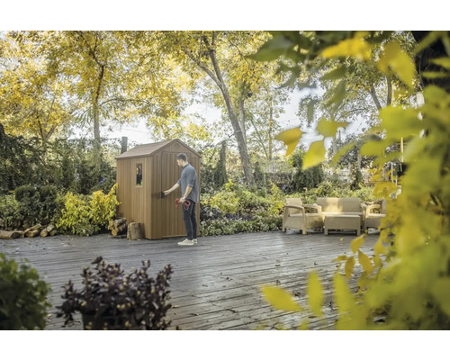 Muž otvára záhradný domček obklopený stromami a záhradným nábytkom.