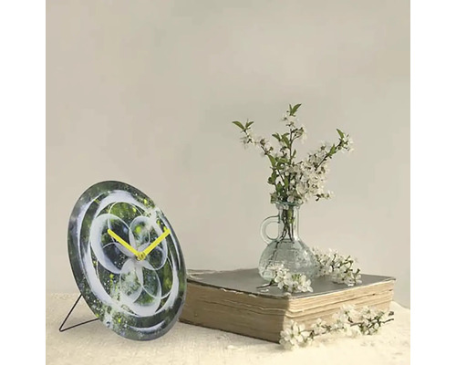 Dekoratívne stolové hodiny s abstraktným dizajnom a žltými ručičkami vedľa knihy a vázy s bielymi kvetmi.