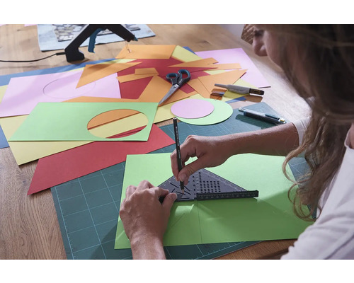 Osoba kreslí uhol a ceruzku na zelený list papiera, obklopený rôznofarebným papierom, nožnicami a rezačkou.