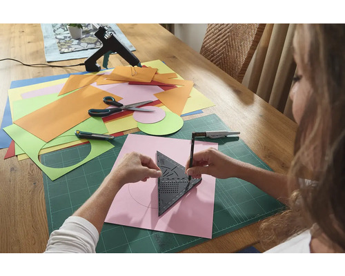 Žena kreslí kruh pomocou trojuholníka s uhlomerom na kus papiera, obklopeného remeselnými materiálmi na stole.