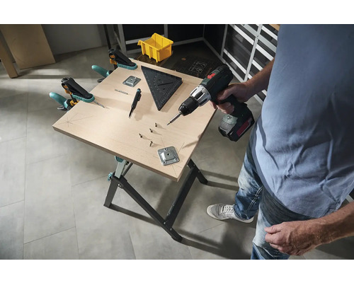 Muž pracuje s akumulátorovým skrutkovačom na pracovnom stole so skrutkami, uholníkom a svorkami.