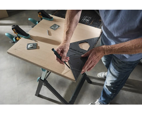 Osoba kreslí uhlomerom a perom čiaru na drevenú dosku, ktorá je pripevnená svorkami a kovovými doskami k pracovnému stolu.