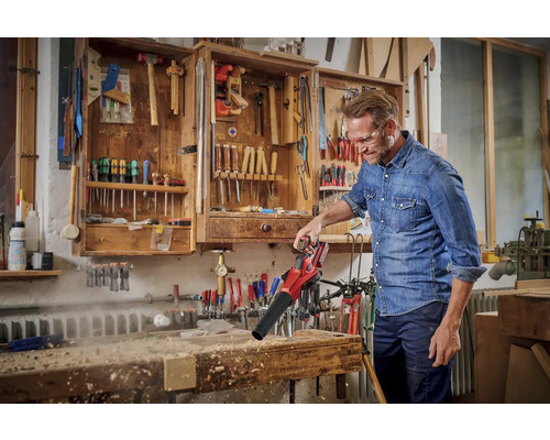 Muž používa akumulátorový fúkač lístia na odfúknutie triesok z pracovného stola. V pozadí náradie v skrini.