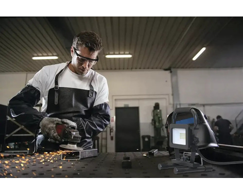 Muž s ochrannými okuliarmi, zásterou a rukavicami brúsi kov pomocou flexy, zatiaľ čo v popredí je staveniskový reflektor.