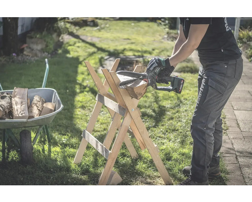 Osoba reže drevo pomocou akumulátorovej reťazovej píly na drevenom kozlíku s kolieskom plným dreva v pozadí.