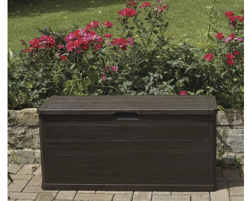 Tmavohnedý záhradný box pred ružovými kríkmi a kamenným múrom.