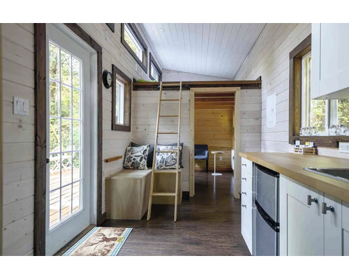 Interiér malého domu s drevenými stenami, rebríkom do vyvýšeného poschodia a kuchynským kútom.