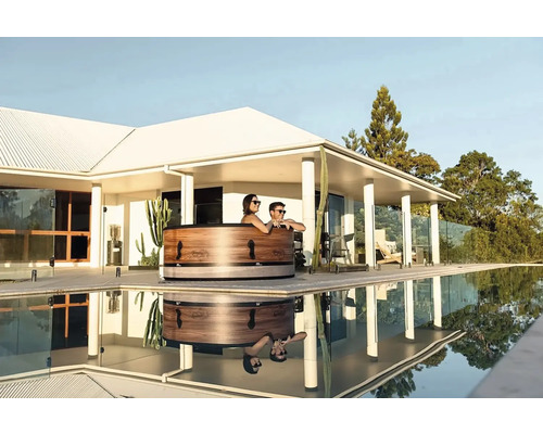 Scéna s drevenou kúpacou kaďou vedľa bazéna, v ktorej sa odrážajú ľudia a okolitá budova.