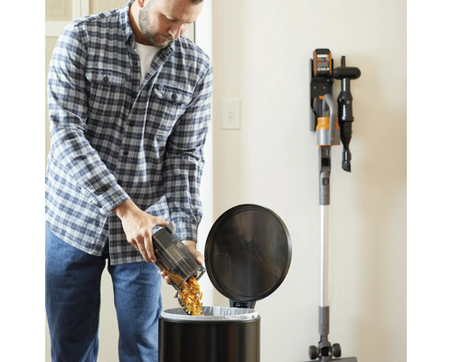 Muž vysypáva nádobu na prach akumulátorového vysávača do odpadkového koša. Na stene je nabíjacia stanica s vysávačom.