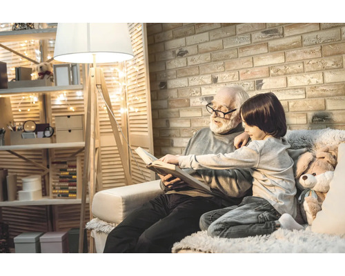 Starý otec číta so svojím vnukom knihu v jasnej, útulnej obývacej izbe s murovanou stenou a stojacou lampou.