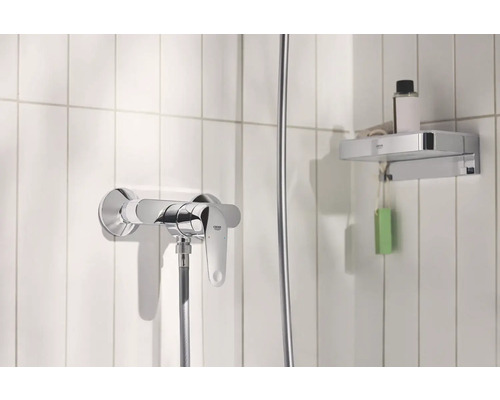 Sprchová batéria a odkladacia polica v kúpeľni s bielymi obkladmi.