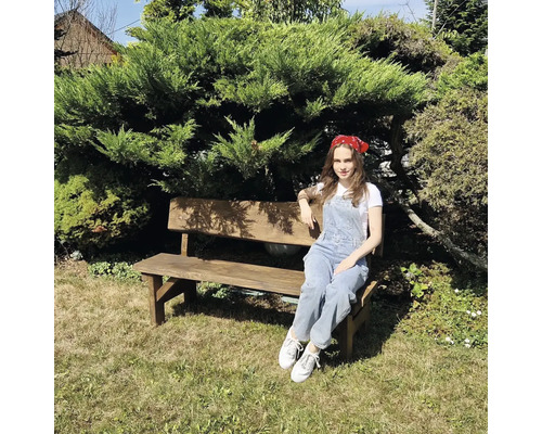 Žena sedí na drevenej záhradnej lavičke pred zeleným kríkom.