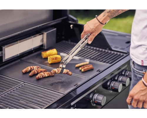 Osoba griluje klobásy, slaninu a kukuričné klasy na grile.