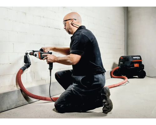 Remeselník vŕta do steny vŕtacím kladivom a odsávaním prachu, pričom je pripravený vysávač