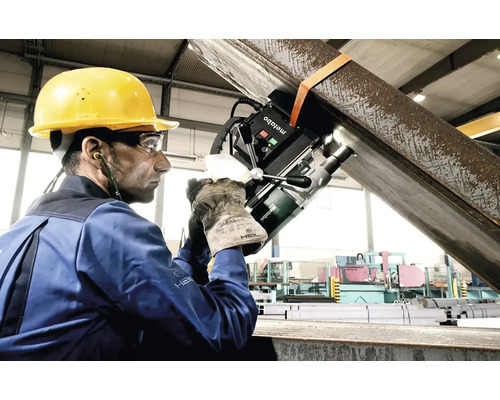 Muž s ochrannou prilbou a okuliarmi obsluhuje magnetickú vŕtačku Metabo na kovovom nosníku v továrnej hale.