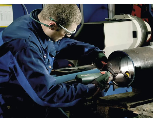 Robotník opracováva kovový obrobok brúskou. Má na sebe pracovný odev a ochranné okuliare.