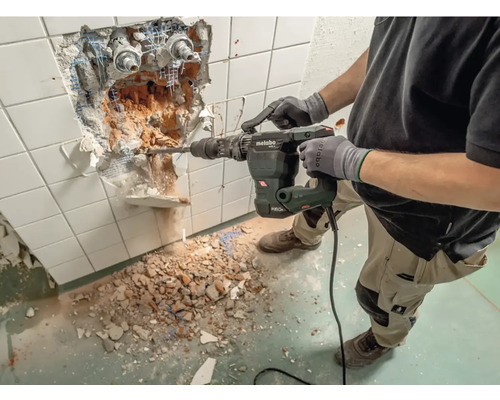 Pracovník používa vŕtacie kladivo na odstránenie dlaždíc zo steny. Na podlahe leží stavebná suť a zvyšky dlaždíc.