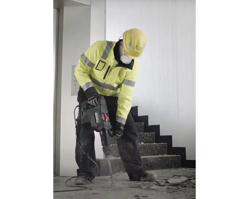 Stavebný robotník s ochrannou prilbou a dýchacou maskou používa zbíjacie kladivo.