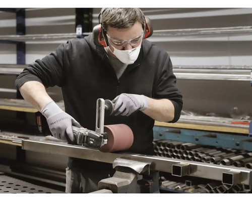 Muž používa brúsku na kovový kus. Nosí ochranný odev.