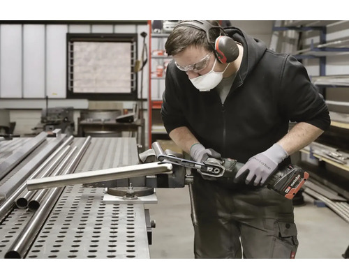 Muž brúsi kovovú rúrku pásovou brúskou v dielni. Má na sebe ochranné pomôcky ako masku a ochranu sluchu.