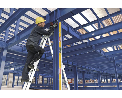 Stavebný robotník s helmou a vodováhou na rebríku pracuje na oceľovej konštrukcii.