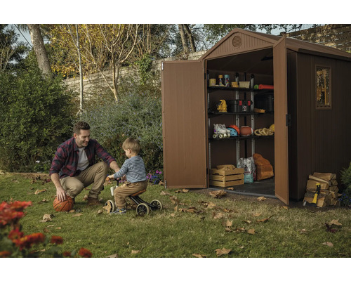 Scéna s plastovým záhradným domčekom s policami, ktorý je plný rôznych vecí, zatiaľ čo sa muž a dieťa hrajú na trávniku.