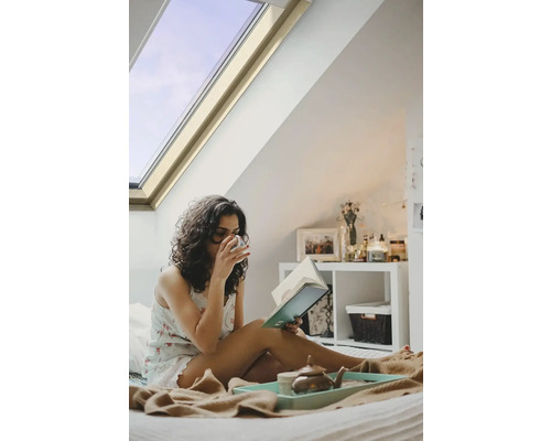 Žena číta knihu v posteli pod strešným oknom.