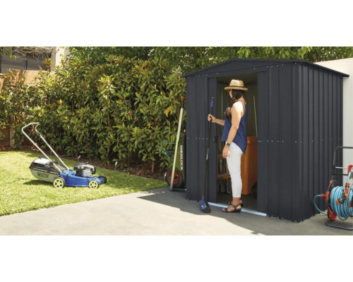 Záhradná scéna s kosačkou na trávu a záhradným domčekom, v ktorom stojí žena