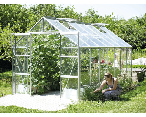 Skleník s otvorenými oknami a rastlinami a žena pracujúca v záhrade.